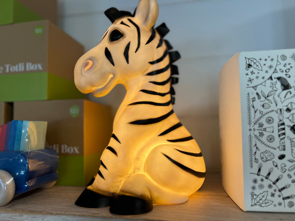 Zebra shaped nightlight facing to the left.  The light is on and shows a warm yellow glow.  The Zebra is in a sitting position.