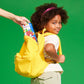 a child wearing a yellow backpack, hert face is turned towards the camera, we can see an adult reaching into the backpack taking out a tin set of microMag magnet tiles