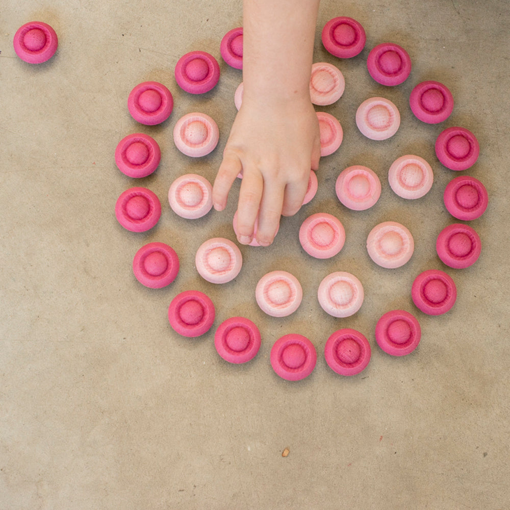 Wooden circle mandala pieces in various shades of pink are arranged in a circle pattern.  A small childs hand is picking up a piece from the centre.