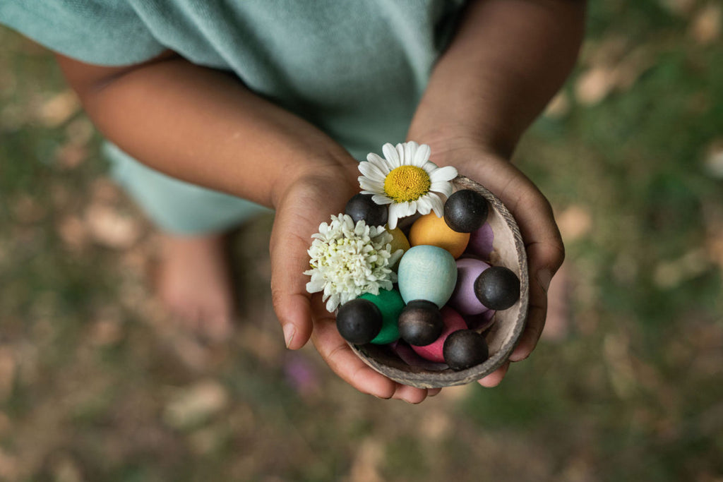 a small child holds a half cocont shell containing flowers and small wooden baby nins